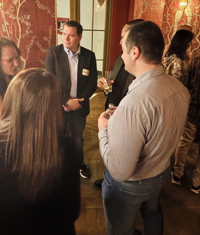 Two men in conversation at a cocktail party, one wearing a name tag and holding a drink, ornate red wallpaper behind them