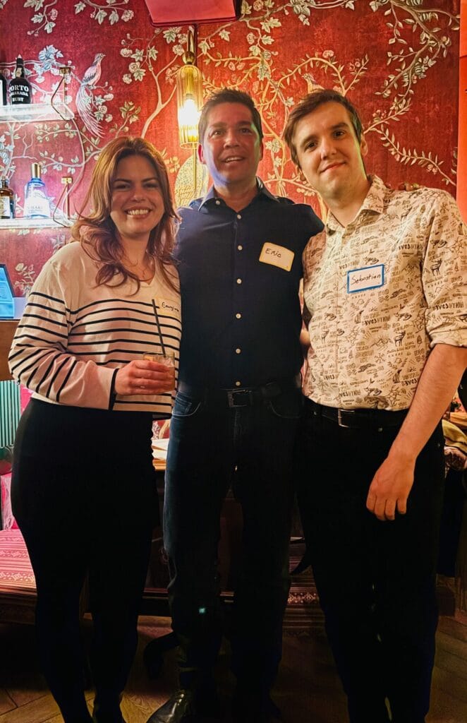 Eric Melchor wearing an Eric name tag posing with two guests at a cocktail party in front of red floral wallpaper