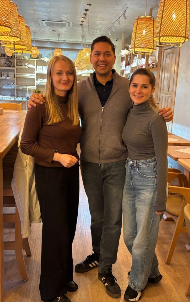 Eric Melchor posing with two women inside a warmly lit cafe with woven pendant lights overhead